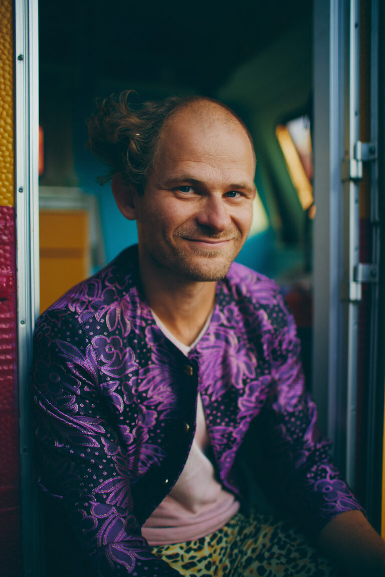 Oskar Rask smiling at the camera. He is sitting in his caravan doorway with a fun one-sided hairstyle and wearing a purple fancy jacket with leopard tights.