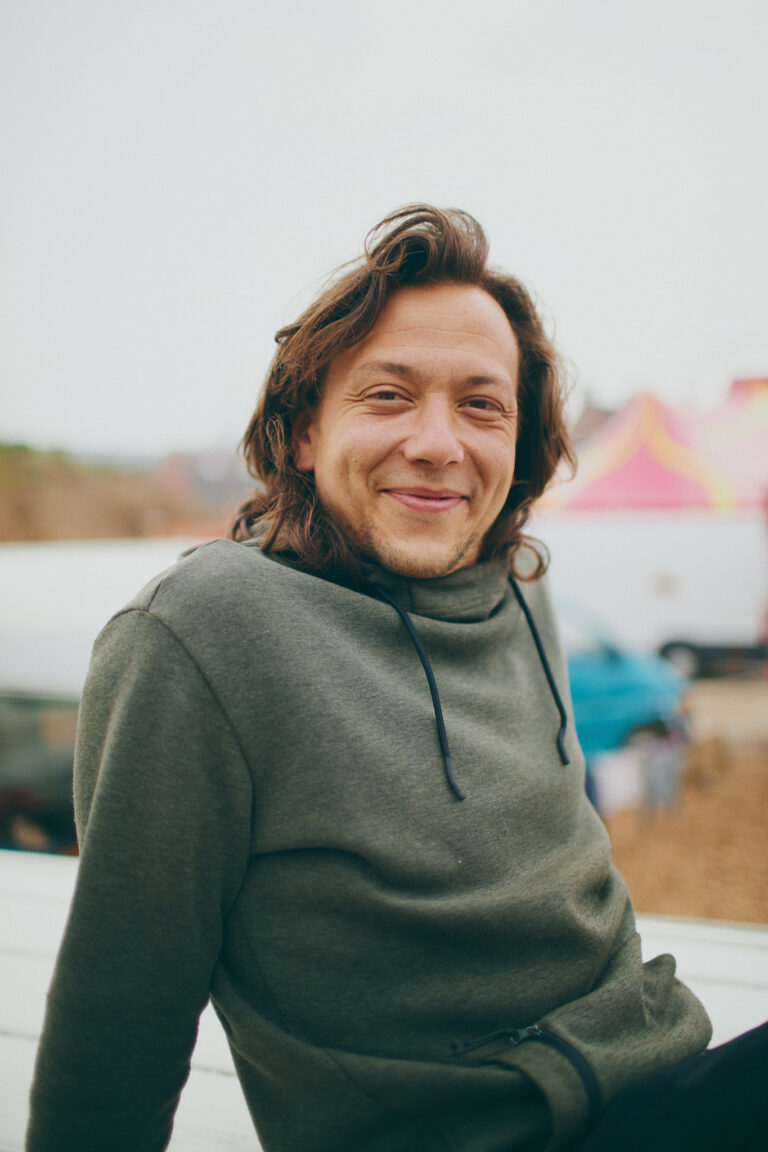 Thibaud is sitting in the caravan camp on top of his van wearing a cosy hoodie and smiling happily.