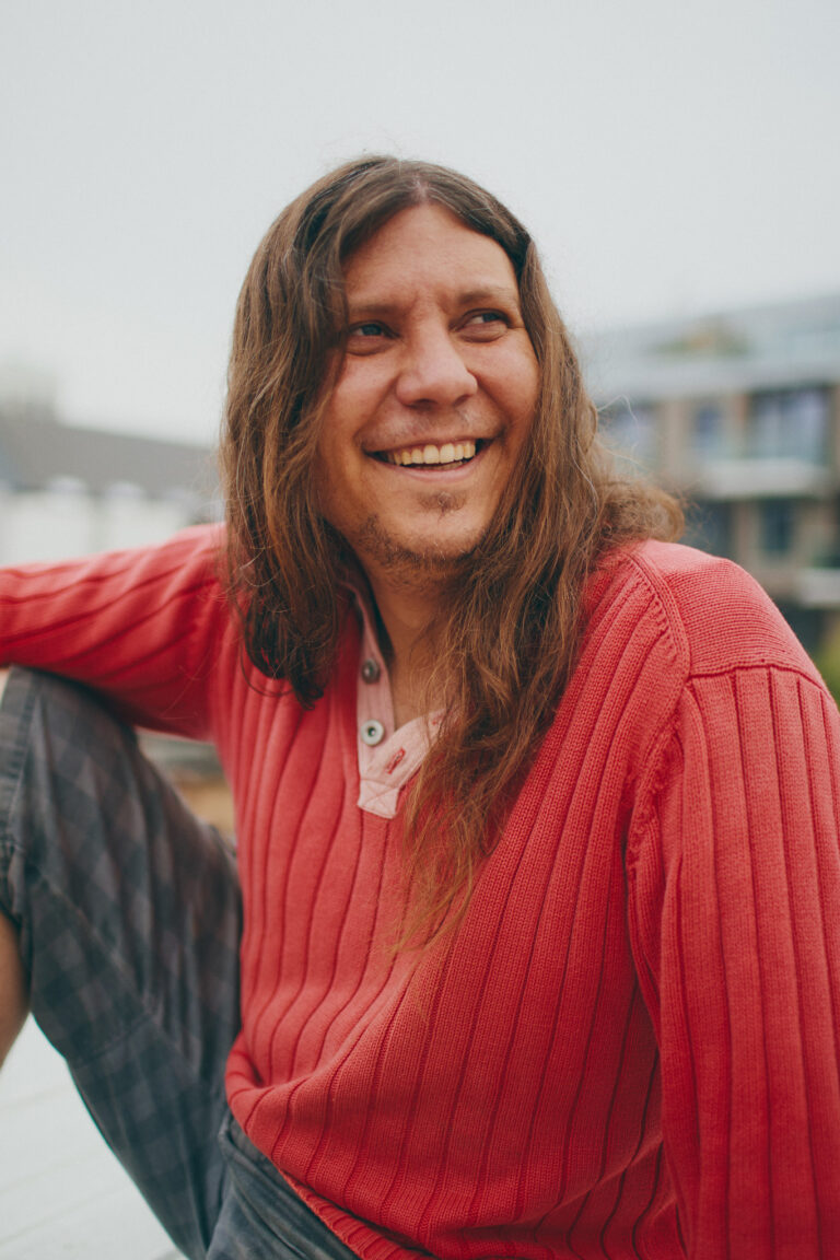 Carlos Ferrer wearing a red pullover is looking past the camera with a happy smile on his face and his long hair open on a cloudy day.