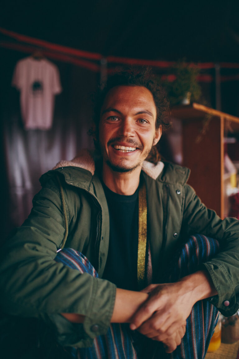 Davide Arra photographed in the small popcorn tent before the performance. Davide is wearing stripy pants, a winter coat and smiling at the camera