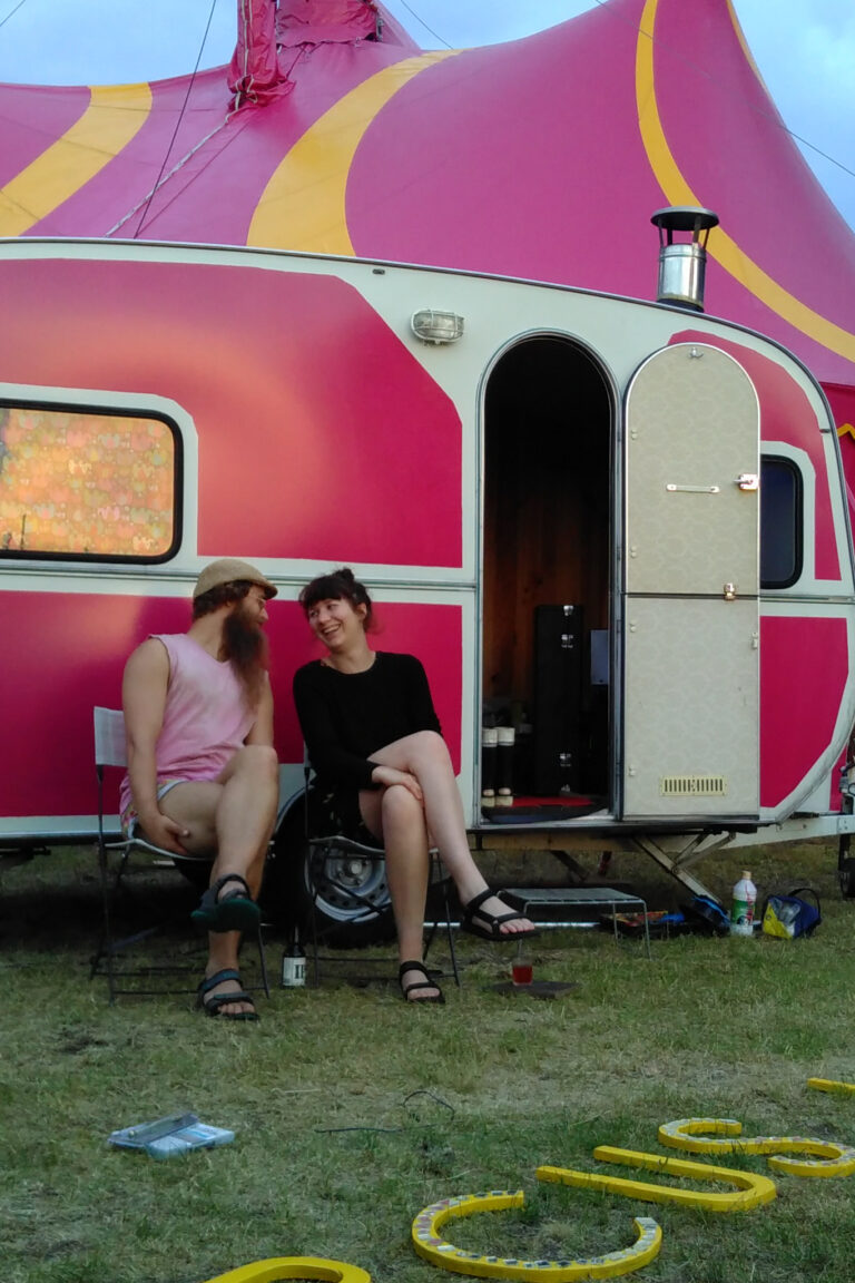 Rhianne De Beer and Oskar Rask are laughing with each other in front of the pink tent and caravan in the eveningtime