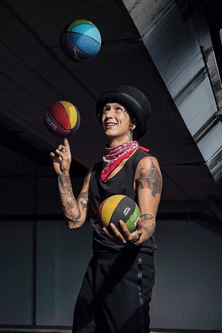 Oscar Quevedo is juggling some colorful basketballs outside. He is dressed in black, wearing a stylish hat and his tattooed arms are bare, showing a large circus tent tattoo on his bicep.